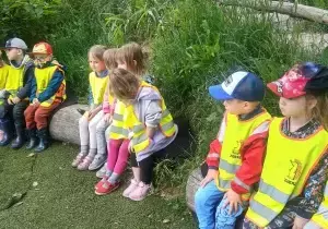 Dzieci siedzą w lasku. Mają na sobie kamizelki odblaskowe. Siedzą na długich pieńkach