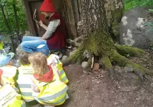 Dzieci siedzą w lasku. Mają na sobie kamizelki odblaskowe. Przed nimi stoi pan przebrany za krasnala.