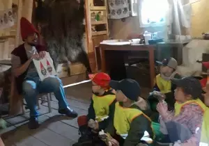 Dzieci siedza w pomieszczeniu. Mają na sobie kamizelki odblaskowe a na głowach czapki. Przed nimi siedzi pan przebrany za krasnala.