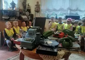 Dzieci siedzą na ławkach. Na środku stoi okrągły st&oacute;ł.