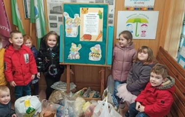 Dzieci stoją przy tablicy z plakatem informacyjnym. Uśmiechaja się do zdjęcia. Przed nimi stoi zebrana karma dla zwierząt leśnych.