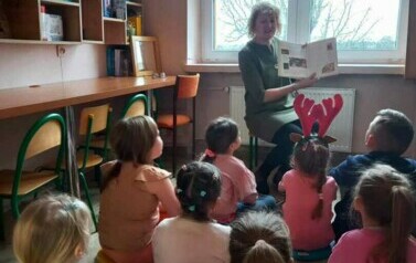 Nauczycielka siedzi na krzesełku. Trzyma w ręku książkę. Dzieci siedzą na podłodze. Patrzą na nauczycielkę.