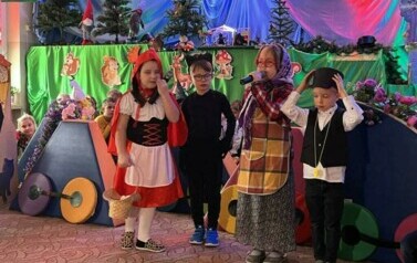 Dziewczynka przebrana za czerwonego kapturka stoi na środku. Obok chłopiec przebrany za wilka. Dalej dziewczynka przebrana za babcię m&oacute;wi przez mikrofon. Obok stoi chłopiec przebrany za zawiadowcę.