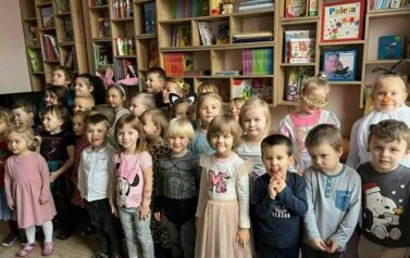 Dzieci stoją i uśmiechają się do zdjęcia. Są w bibliotece przedszkolnej.