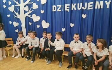 Dzieci siedzą na krzesełkach, uśmiechają się.