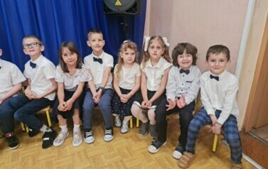 Dzieci siedzą na krzesełkach, uśmiechają się.