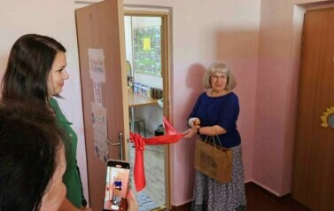 Pani Bibliotekarka przecina nożyczkami czerwoną wstęgę. Wstęga zawieszona jest przed drzwiami biblioteki przedszkolnej.
