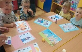 Dzieci siedzą przy stolikach. Układają puzzle. Na głowach mają opsaki z napisem dzień postaci z bajek.