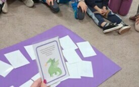 Dzieci siedza na dywanie. Przed nimi leży fioletowy karton. Nauczycielka siedzi przed nimi. Trzyma w ręku kartkę z zagadką.