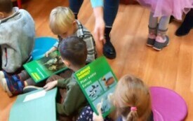 Dzieci siedzą w bibliotece na kolorowych pufach. Pani bibliotekarka wyciąga rękę do dziecka. Dzieci trzymają w rekach książki.