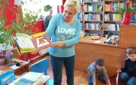 Pani bibliotekarka trzyma w ręku książkę o tygrysku. Czyta ją dzieciom Stoi. Na przeciwko niej siedzą dzieci na kolorowych pufach.