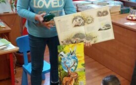 Pani bibliotekarka trzyma w ręku otwartą książkę. Stoi przed dziećmi. Dzieci siedzą na kolorowych pufach.