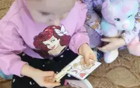 Dziewczynka siedzi na dywanie. Uśmiecha się. Przypina spinacz do kartki. Na kartce jest wizerunek misia. Obok niego są trzy kształty. Dziewczynka siedzi obok. Przygląda się.