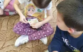 Dziewczynka siedzi na dywanie. Uśmiecha się. Przypina spinacz do kartki. Na kartce jest wizerunek misia. Obok niego są trzy kształty. Obok siedzi chłopiec. Przygląda się.