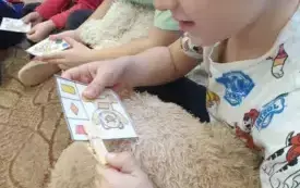 Chłopiec siedzi na dywanie. Uśmiecha się. Przypina spinacz do kartki. Na kartce jest wizerunek misia. Obok niego są trzy kształty.