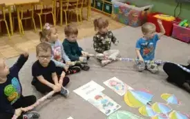 Dzieci siedzą na dywanie. W rękach trzymają gumę. Przed nimi leżą kartki z prawami dziecka i kolorowe parasolki wycięte z papieru.