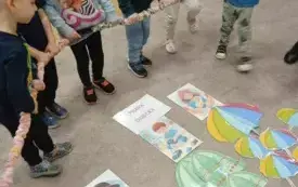 Dzieci stoją na dywanie. W rękach trzymają gumę. Przed nimi leżą kartki z prawami dziecka i kolorowe parasolki wycięte z papieru.