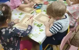 Dzieci siedzą przy stolikach. Na stolikach leżą ceraty. Dzieci trzymają w rękach pędzelki. Na środku stołu stoją miseczki z kolorową kaszą i klejem