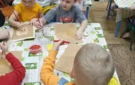 Dzieci siedzą przy stolikach. Na stolikach leżą ceraty. Dzieci trzymają w rękach pędzelki. Na środku stołu stoją miseczki z kolorową kaszą i klejem