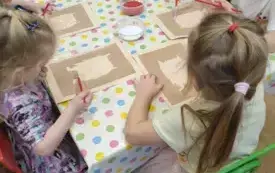 Dzieci siedzą przy stolikach. Na stolikach leżą ceraty. Dzieci trzymają w rękach pędzelki. Na środku stołu stoją miseczki z kolorową kaszą i klejem.
