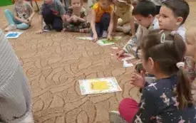 Dzieci siedzą na dywanie. Przed nimi leżą kartki z kolorowymi stworkami.