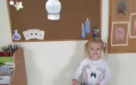 Dziewczynka stoi przy tablicy przedszkolnej. Na tablicy jest napis andrzejki. Dziewczynka trzyma w ręku kartkę.