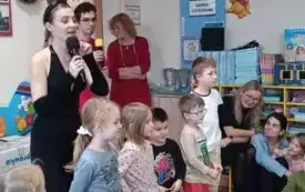 Pani stoi z mikrofonem. Jest przebrana za kota. Dzieci stoją przed nią. Jeden obok drugiego. 