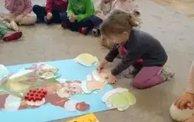 Dziecisiedzą na dywanie. Układają czapkę na głowie mikołaja. Mikołaj wycięty jest z kartonu. Leży na dywanie.