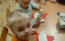 Dzieci siedzą przy stoliku. Robią prace plastyczną. Przyklejają elementy mikołaja.