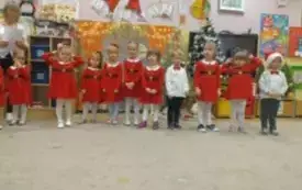 Dzieci stoją jeden obok drugiego w sali przedszkolnej. Dziewczynki ubrane są w czerwone sukienki. Chłopcy w odświęten koszule. 