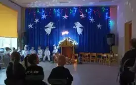 Zdjęcie żł&oacute;bka. Obok niego siedzą dzieci ubrane odświętnie. Nad żł&oacute;bkiem wiszą białe anioły wycięte z kartonu. 