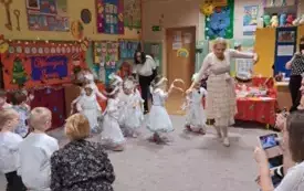 Dziewczynki ubrane w śnieżynki tańczą w sali przedszkolnej. Razem z nimi tańczy nauczycielka ubrana na biało.