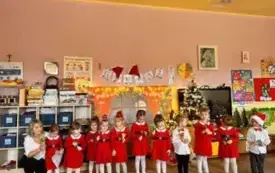 Dzieci stoją w sali przedszkolnej. Jeden obok drugiego. Dziewczynki ubrane są w czerwone sukienki. Chłopcy odświętnie.