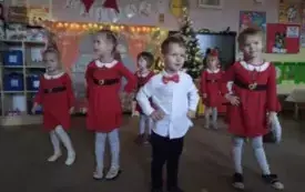 Dzieci stoją w sali przedszkolnej. Dziewczynki ubrane są w czerwone sukienki. Na przodzie stoi chłopiec w białej koszuli. Odświętnie ubrany.