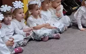 Dziewczynki siedzą na dywanie. Przebrane są za śnieżynki. Na głowach mają opaski z gwiazdkami śnieżnymi. Ubrane są na biało.