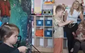 Dziewczynka i chłopiec stoją w sali przedszkolnej. Trzymają w rękach metalowe krążki. Na środku stoi dekroacja iluzjonisty. Z boku widać nauczycielkę.