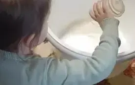 Dziewczynka wsypuje szklankę mąki do miski. Miska stoi na stole. St&oacute;łprzykryty jest ceratą.