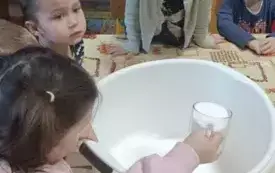 Dziewczynka wsypuje szklankę mąki do miski. Inne dzieci przyglądają się. Miska stoi na stole.