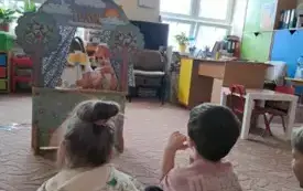 Dzieci siedzą na dywanie. Przed nimi stoi teatrzyk. W teatrzyku są dwie dziewczynki. Trzymają w rękach wykonane figurki na patyczku.