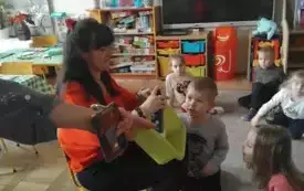 Pani pokazuje dzieciom piankę do golenia. Dzieci siedzą na dywanie. Przyglądają się. 