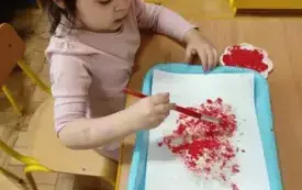Dziewczynka siedzi przy stoliku. Maluje farbami serce. 