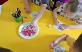 Dzieci siedzą przy stolikach kolorują karteczki z napisem Dzień Dziecka.