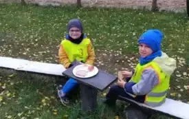 Chłopcy siedzą na ławeczce i jedzą kiełbasę.