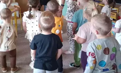 Dzieci tańczą w sali przedszkolnej. Przed nimi stoi pan.